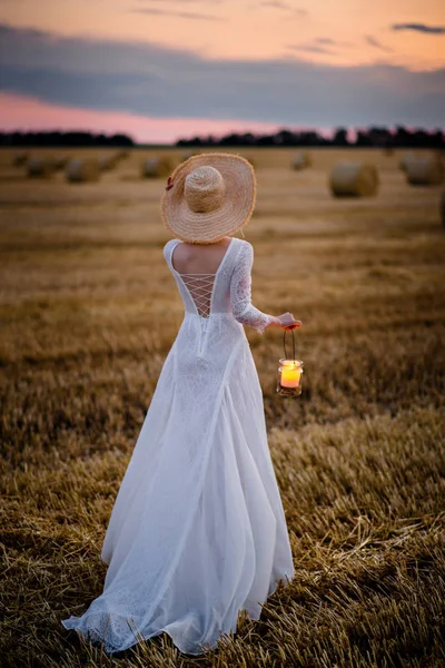 彼女の手にランプを持つ白いドレスを着た女性は、日没後にフィールドを横切って歩きます — ストック写真