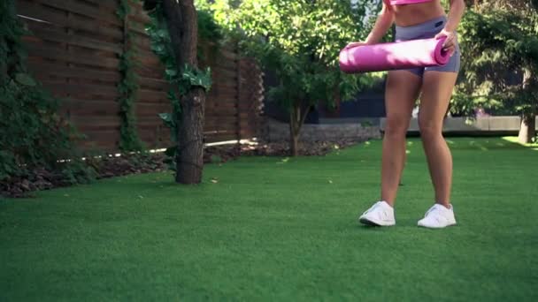 Jeune femme roule tapis de fitness rose sur l'herbe verte, au ralenti — Video