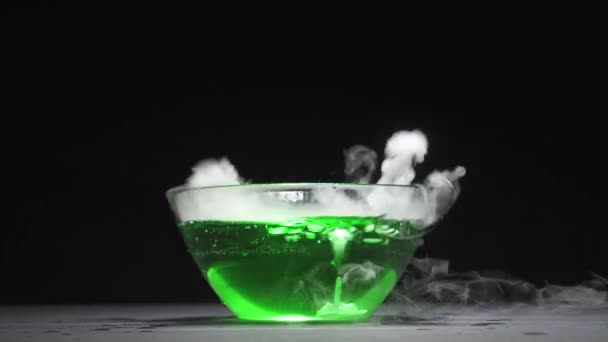 Green liquid boils giving off smoke in a glass bowl on a black background — Stock Video