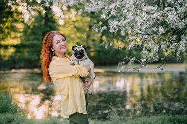 Genç kızıl saçlı bir kadın köpeğini önünde tutuyor. Sahibi ve evcil hayvanı dışarıda eğleniyorlar.. 