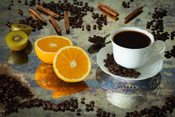 a cup of black coffee with spilled coffee beans, pieces of orange, sticks of cinnamon and kiwi.  Photo in vintage style