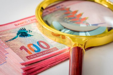 Üzerinde büyüteç olan 100 Bolivya banknotundan oluşan bir deste. Para birimi dalgalanmaları ve zor ekonomik durum kavramı