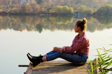 Mavi kot giyen, kırmızı gömlekli ve siyah çizmeli genç bir kız gölün kenarındaki ahşap köprüde oturuyor. Dışarıda oturan genç bir kız. İnsanlar, doğa, seyahat kavramı.