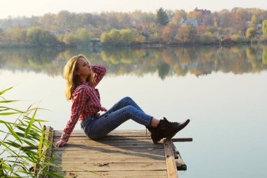Mavi kot giyen, kırmızı gömlekli ve siyah çizmeli genç bir kız gölün kenarındaki ahşap köprüde oturuyor. Dışarıda oturan genç bir kız. İnsanlar, doğa, seyahat kavramı.