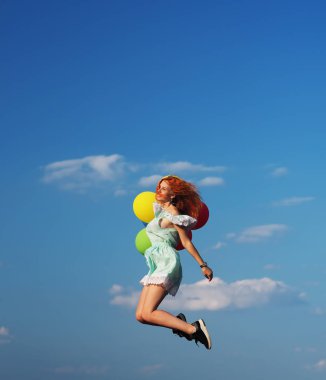 Mavi gökyüzünde zıplayan renkli balonları olan genç kızıl saçlı kız.