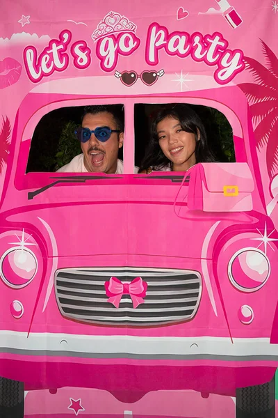 İki arkadaş pembe bir arabanın arkasında poz verirken eğleniyorlar. Üzerinde parti yazılıydı. Barbie temalı bir partinin tadını çıkarıyorlardı.