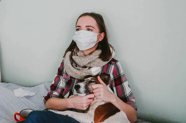 A young woman in a medical mask and petting a dog suffers from allergies at home — Stock Photo, Image