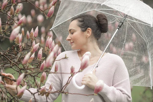春白いレースのドレスと透明な傘で咲く木の近くに立っているジーンズで美しいロマンチックな赤髪の女の子。カメラを見て雨の下で道路の近くの若いモデル. — ストック写真
