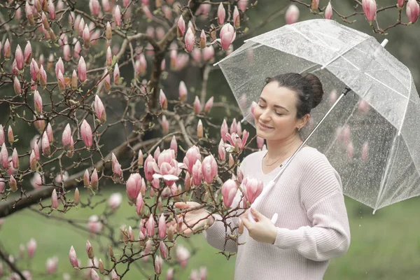 Wiosna piękne romantyczne czerwone włosy dziewczyna w białej sukni koronki i dżinsy stoi w pobliżu kwitnącego drzewa z przezroczystym parasolem. Młody model w pobliżu drogi pod deszcz patrząc na aparat fotograficzny. — Zdjęcie stockowe