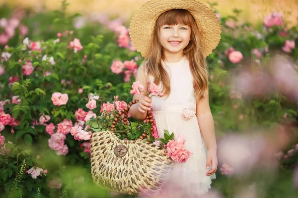 Ein kleines Mädchen mit schönen langen blonden Haaren, in einem hellen Kleid und einem Kranz aus echten Blumen auf dem Kopf, im Garten einer Teerose — Stockfoto