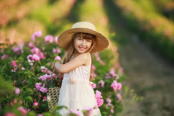 A little girl with beautiful long blond hair, dressed in a light dress and a wreath of real flowers on her head, in the garden of a tea rose — Stock Photo, Image