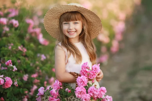 Egy kislány, gyönyörű, hosszú szőke hajú, öltözött egy könnyű ruhát, és a koszorú valódi virágok a fején, a kertben egy tea rózsa — Stock Fotó