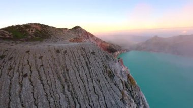 Gündoğumunda, Endonezya 'nın Doğu Java kentindeki turkuaz sülfür gölüne tepeden bakan engebeli Ijen Volkanı' nın engebeli krater kenarının üzerinde sinematik FPV insansız hava aracı uçuşu. Göz kamaştırıcı volkanik manzara ve sabah ışığı.