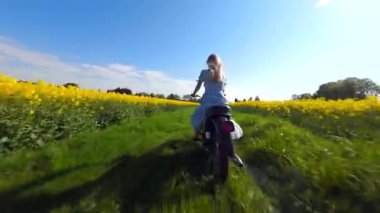 Güneşli bir günde sarı kolza tohumu tarlasında motosiklet süren bir kadının sinematik FPV görüntüsü. Özgürlük ve yaz temaları için enerjik kırsal yaşam tarzı manzarası.