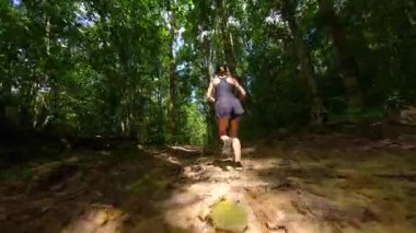 Aktif giyimli kadın koşucunun tropikal bitki örtüsüyle çevrili yaprak orman yolunda koşarken çekilmiş sinematik FPV görüntüsü. Doğa ve macera temaları için ideal.