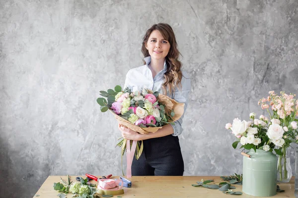 Jeune femme regardant la caméra, tient un bouquet luxueux dans ses mains. fleuriste créer beau bouquet dans la boutique de fleurs. Assistante ou propriétaire dans un studio de design floral . — Photo