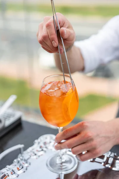 Бармен делает коктейль Aperol spritz. Ошибочное стекло, избирательная фокусировка. Алкогольный напиток на основе барной стойки со льдом и апельсинами. Вечеринка на открытом воздухе — стоковое фото