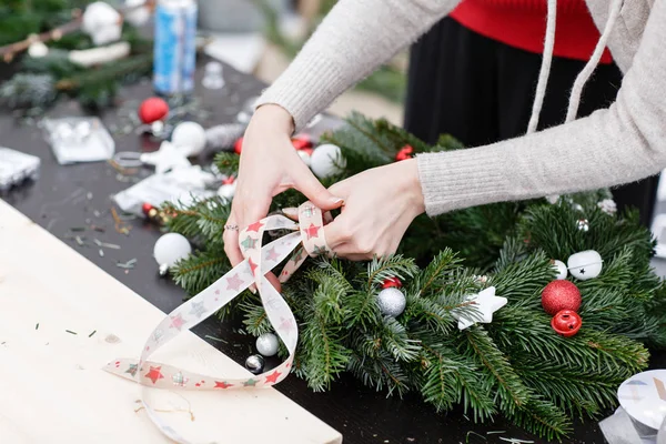 Mujer atando una cinta de lazo, decorado una corona de Navidad. Adjunta juguetes y decoración con pistola de pegamento. Manos de cerca. Clase magistral sobre la fabricación de ornamentos decorativos . — Foto de Stock