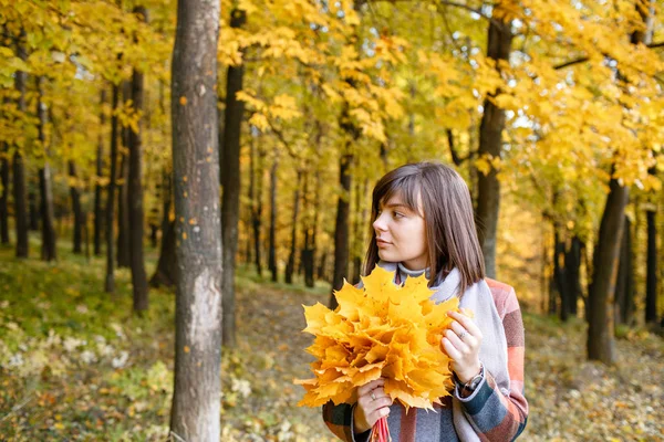 Portrait de jeune femme de mode en plein air. Brunette femme dans le parc d'automne avec manteau à carreaux à la mode et écharpe. Exploitation de feuilles jaunes — Photo