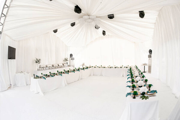 Interior of tent for wedding diner, ready for guests. Served round banquet table outdoor in marquee decorated hydrangea flowers, Golden dishes and green napkins. Catering concept