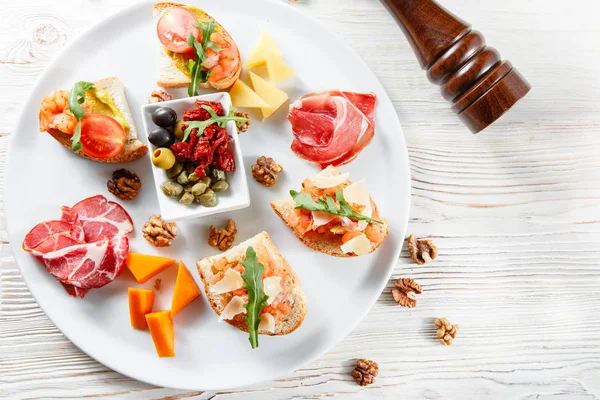 Plato con aperitivos italianos. Bruschetta con tomates cherry y camarones. Queso parmesano, jamón, alcaparras verdes, aceitunas, tomates secados al sol y nueces . —  Fotos de Stock