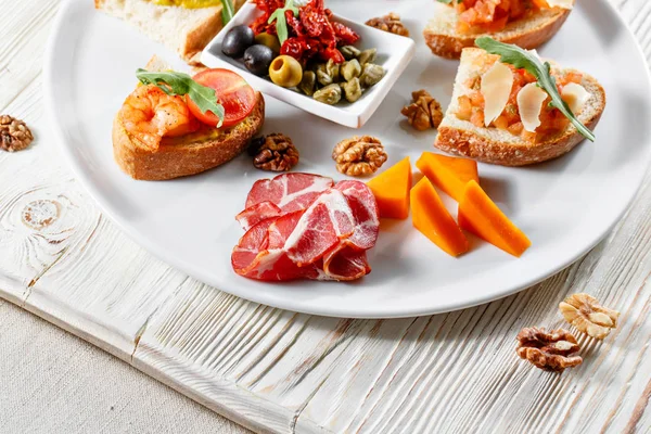 Plato con aperitivos italianos. Bruschetta con tomates cherry y camarones. Queso parmesano, jamón, alcaparras verdes, aceitunas, tomates secados al sol y nueces . — Foto de Stock