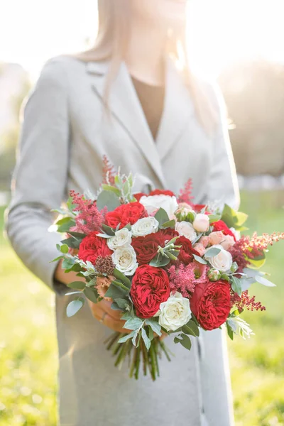 Прекрасна жінка тримає гарний осінній весільний букет. квіткова композиція з білими і червоними садовими трояндами. Зелений газон на фоні. Яскравий світанок або захід сонця — стокове фото