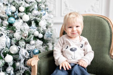 Portre küçük çocuk. Yeni Yılınız Kutlu Olsun. süslü Noel ağacı. Noel sabahı parlak oturma odasında. yeşil sandalye üzerinde oturan