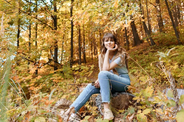 Porträt einer jungen Frau im Herbstpark, die auf einem Holzstamm sitzt. Bäume mit gelbem Laub im Hintergrund, schönes Sonnenuntergangslicht. warmer Pullover auf den Schultern — Stockfoto