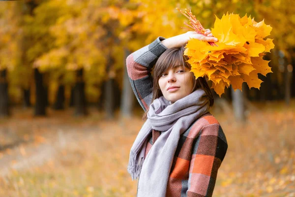 Portrait de jeune fille en plein air. Bouquet avec des feuilles jaunes dans les mains. Brunette femme dans le parc d'automne avec manteau à carreaux à la mode et écharpe . — Photo