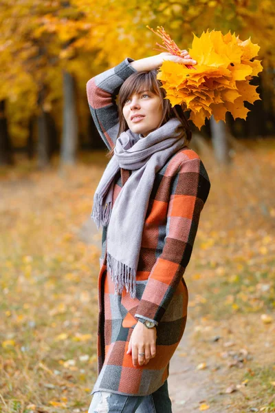 Retrato de menina jovem ao ar livre. Buquê com folhas amarelas nas mãos. Mulher morena no parque de outono com casaco xadrez elegante e cachecol . — Fotografia de Stock
