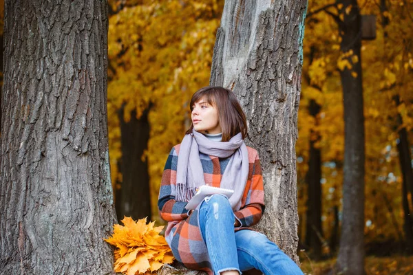 University Park, concepto de estudio. Retrato de mujer joven en bosque otoñal. Escribe en un cuaderno. Mujer morena en el parque de otoño con abrigo a cuadros de moda y bufanda . —  Fotos de Stock