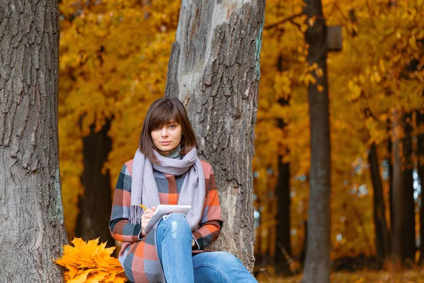 University Park, concept d'étude. Portrait de jeune femme dans la forêt d'automne. Écrit dans un carnet. Brunette femme dans le parc d'automne avec manteau à carreaux à la mode et écharpe . — Photo