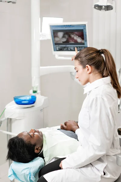 Urocze, piękna dziewczyna lekarz dentysta. Młody Afroamerykanin pacjent w fotelu w klinice stomatologicznej. Medycyna, zdrowie, Stomatologia koncepcja. dentysta przeprowadza inspekcję i stwierdza — Zdjęcie stockowe