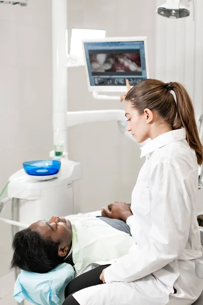 Encantadora doctora dentista hermosa. Joven paciente afroamericano en silla en la clínica dental. Medicina, salud, concepto estomatológico. dentista realiza inspección y concluye — Foto de Stock