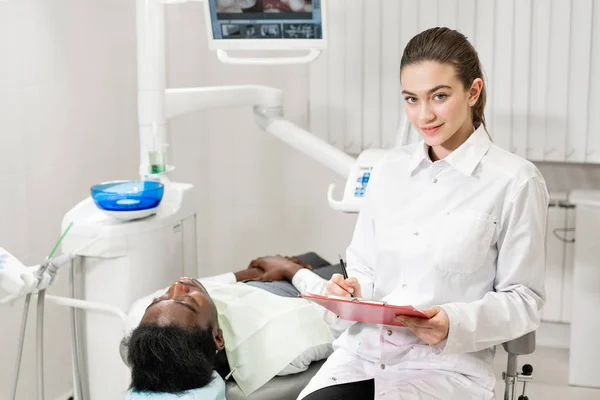 Encantadora doctora dentista hermosa. Joven paciente afroamericano en silla en la clínica dental. Medicina, salud, concepto estomatológico. dentista lleva a cabo la inspección y concluye. Mujer sonriendo — Foto de Stock