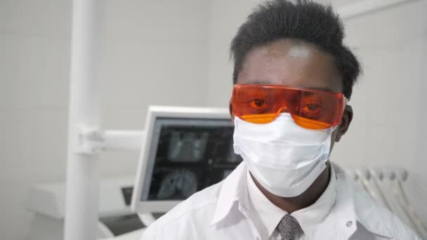 Βάζει σε ένα ιατρικό πορτοκαλί γυαλιά ασφαλείας. Νέοι Αφρικανικός Αμερικανός οδοντίατρος αρσενικό ο γιατρός στην ιατρική μάσκα. Ιατρική, υγεία, Στοματολογία έννοια. οδοντίατρο που διεξάγει επιθεώρηση και καταλήγει στο συμπέρασμα — Αρχείο Βίντεο