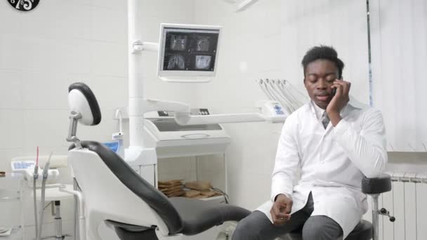 Νέοι αφρικανική αμερικανική αρσενικό ο γιατρός οδοντίατρος στο οδοντιατρείο. Μιλώντας για τον ασθενή στο τηλέφωνο. Ιατρική, υγεία, Στοματολογία έννοια. — Αρχείο Βίντεο