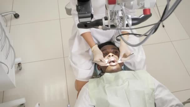 La doctora dentista usa un microscopio. Joven paciente afroamericano en silla en la clínica dental. Medicina, salud, concepto estomatológico. dentista realiza inspección y concluye . — Vídeos de Stock