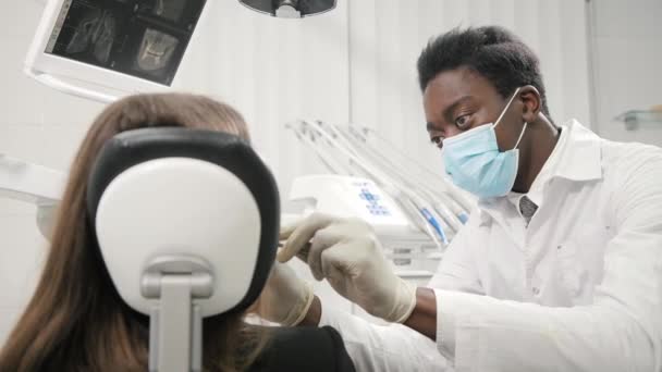Junger afrikanisch-amerikanischer Zahnarzt mit medizinischer Maske. schöne Patientin am Stuhl in der Zahnklinik. Medizin, Gesundheit, Stomatologie-Konzept. Zahnarzt führt Inspektion durch und schließt — Stockvideo