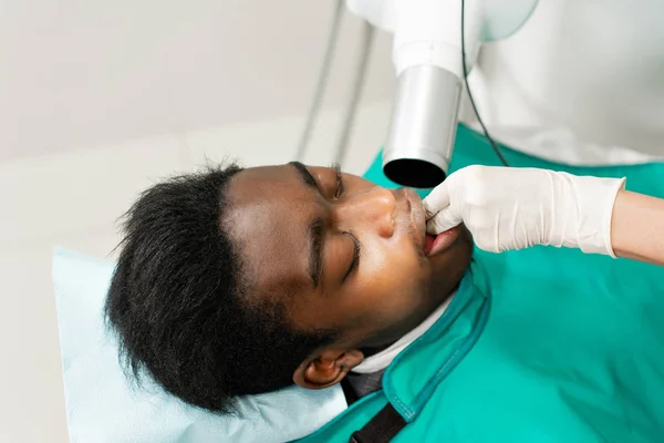 Mujer dentista usando máquina de rayos X, paciente acostada en silla en odontología. Joven hombre afroamericano con mal diente. Medicina, salud, concepto de estomatología . — Foto de Stock