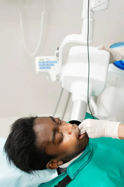 Mujer dentista usando máquina de rayos X, paciente acostada en silla en odontología. Joven hombre afroamericano con mal diente. Medicina, salud, concepto de estomatología . — Foto de Stock