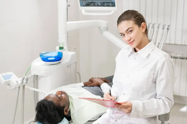 Okouzlující krásná dívka zubař hledí do kamery. Mladý Američan Afričana pacient v křesle na zubní klinice. Lékařství, zdravotnictví, stomatologie koncept. zubní lékař provádí kontroly a uzavírá. — Stock fotografie