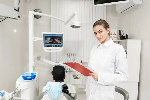 Encantadora hermosa chica dentista mira a la cámara. Joven paciente afroamericano en silla en la clínica dental. Medicina, salud, concepto estomatológico. dentista realiza inspección y concluye . — Foto de Stock