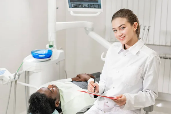 Encantadora hermosa chica dentista mira a la cámara. Joven paciente afroamericano en silla en la clínica dental. Medicina, salud, concepto estomatológico. dentista realiza inspección y concluye . — Foto de Stock