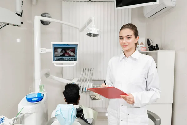 Encantadora hermosa chica dentista mira a la cámara. Joven paciente afroamericano en silla en la clínica dental. Medicina, salud, concepto estomatológico. dentista realiza inspección y concluye . — Foto de Stock