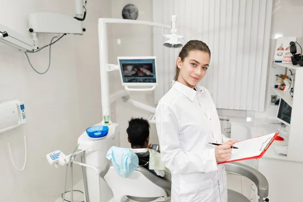Okouzlující krásná dívka zubař hledí do kamery. Mladý Američan Afričana pacient v křesle na zubní klinice. Lékařství, zdravotnictví, stomatologie koncept. zubní lékař provádí kontroly a uzavírá. — Stock fotografie