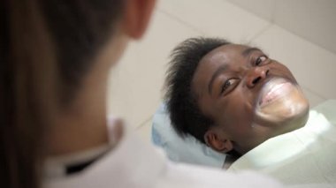 Güzel bir kadın doktor diş hekimi. Genç Afro-Amerikan erkek hasta sandalyeye diş kliniği. Tıp, sağlık, stomatology kavramı. diş hekimi muayene yapar ve bitirmek adl. kameraya bakarak
