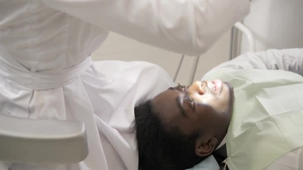 女性医師歯科医は顕微鏡を使用します。歯科医院の椅子に男性患者は若いアフリカ系アメリカ人。医学、健康、口腔病学の概念。歯科医が検査し、最後. — ストック動画