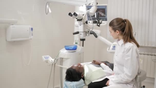 The dentist directs modern microscope to the patient. Beautiful woman doctor dentist. Young African American male patient at chair at dental clinic. Medicine, health, stomatology concept. — Stock Video